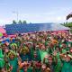 A large group of children wearing matching green clothing and colorful flower garlands raise their hands while standing outdoors. Behind the group is a structure with a rooftop covered in solar panels. The scene is bright and tropical, with palm trees, green grass, and the ocean visible in the background.