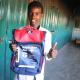A young somalian boy holding a blue and red backpack with an Australian Aid logo in his right hand and his left hand with his thumb up.