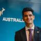 Ricky standing in front of an 'Australia' nation brand banner, dressed in a dark blue suit, white business shirt, and a maroon tie with white chevron pattern. 