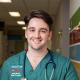 Man standing in a hallway in hospital scrubs with a stethoscope around his neck.