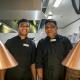 Two men dressed in chef’s uniform, in a kitchen, with heat lamps in the foreground.