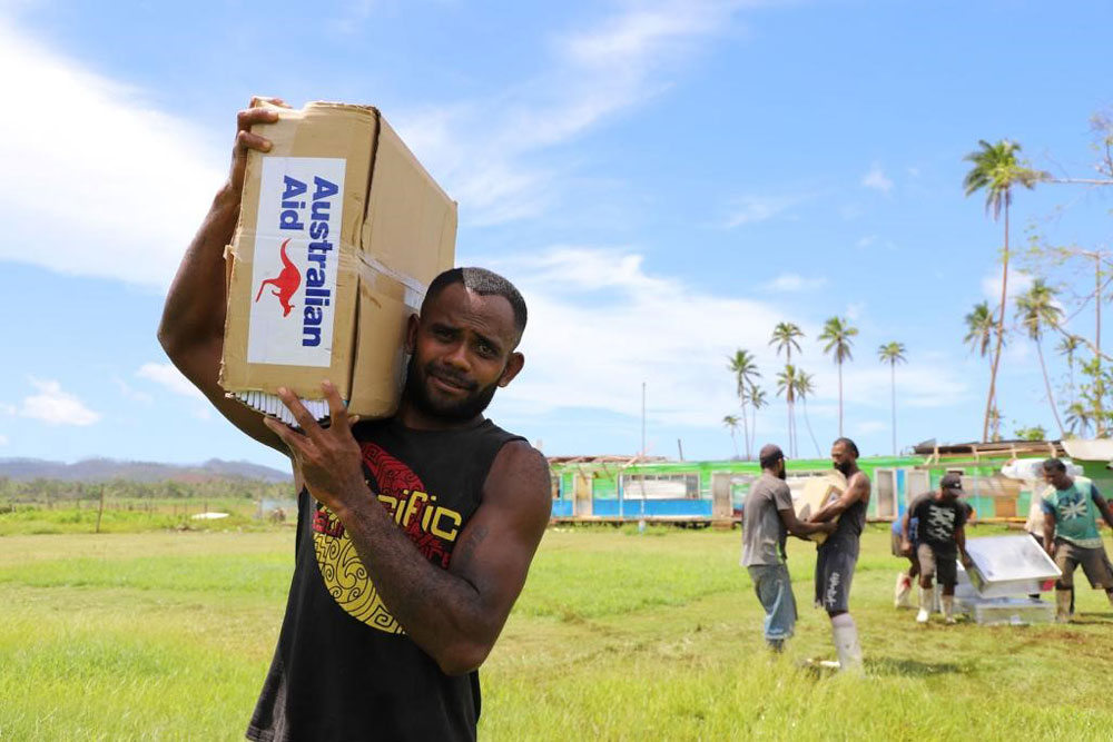 A man carries a box marked ‘Australian Aid’ on his shoulder. He stands in a field with palm trees in the background.