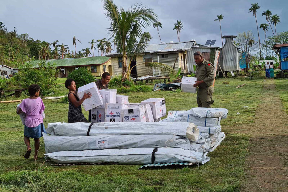 Three locals collect humanitarian relief supplies delivered to a field in Fiji. Buildings and palm trees are in the background.