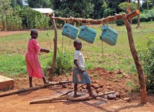 Two girls stand on logs that are connected by rope to containers suspended above the ground. When the logs are pushed down the containers tip and clean water comes out for washing hands.