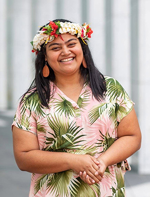 A person standing outdoors wearing a pink shirt with a green tropical leaf pattern. They have long dark hair and are wearing a colorful floral headpiece and orange earrings. Their hands are gently clasped in front of them, and the background is softly blurred vertical panels.