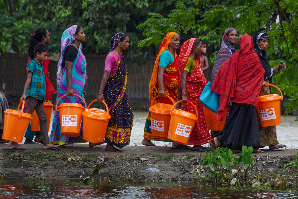 Group of women and children walking with big buckets of dignity kits.
