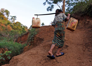 A woman walking up a steep dirt track carrying two containers across her shoulders