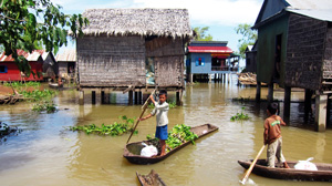 Two boys standing in canoes on muddy water, navigating their way between houses on stilts